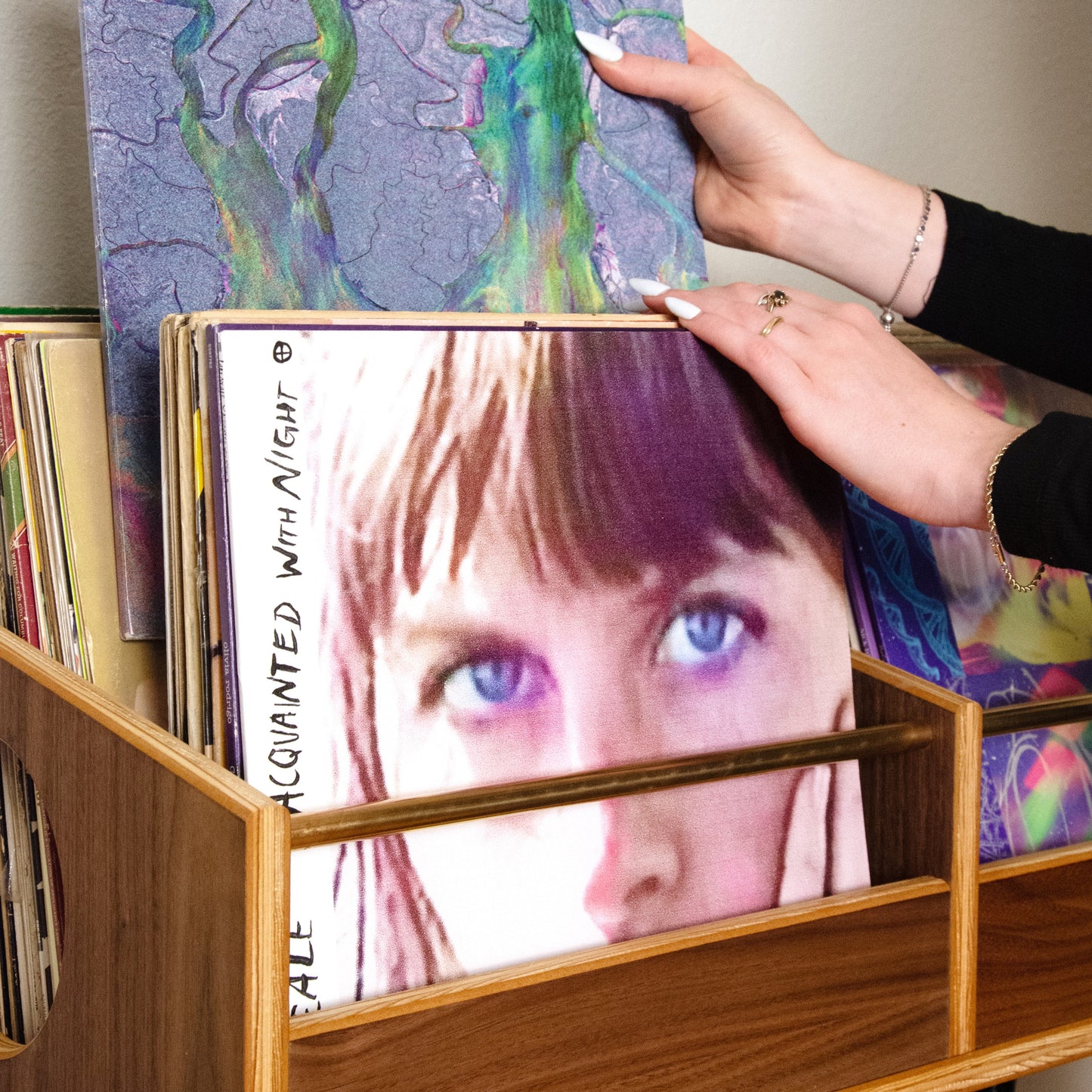 A person's hands holding a vinyl record sleeve with a purple and green marbled pattern, placing it into a wooden record rack filled with other records.