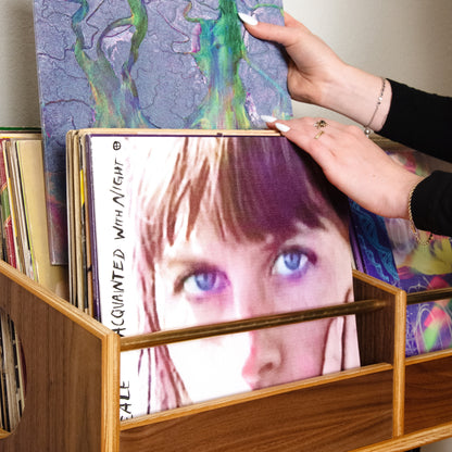 A person's hands holding a vinyl record sleeve with a purple and green marbled pattern, placing it into a wooden record rack filled with other records.
