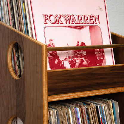 A close-up of a wooden vinyl record shelf filled with records, featuring a prominent red 'FOX WARREN' album cover leaning in the top slot.