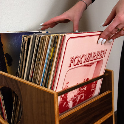 A wooden record rack filled with vinyl records, with a person's hands reaching for a pink 'Fox Warren' record cover against a plain wall.