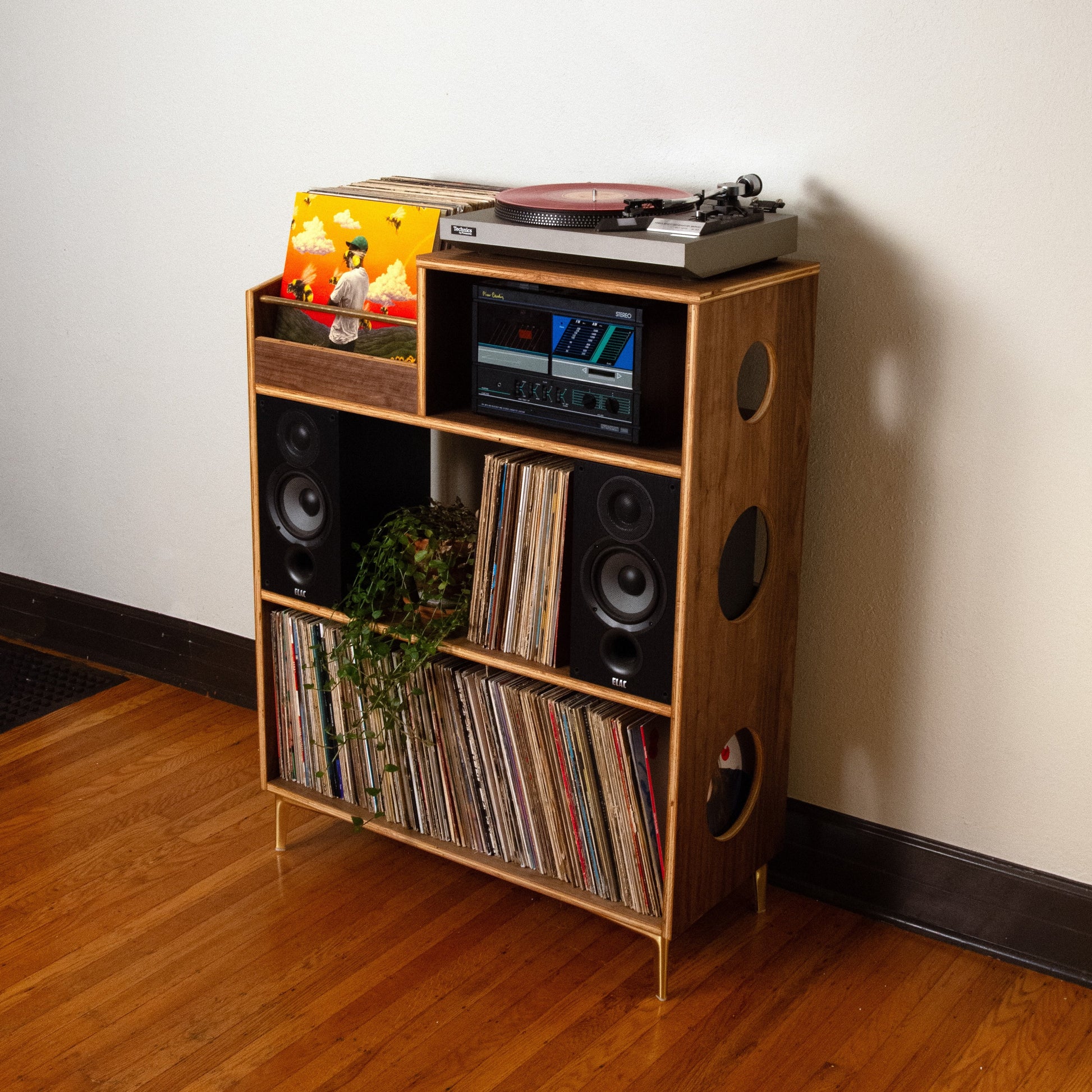 A mid-century modern wooden record player and vinyl record shelf unit, styled with speakers, records, and a turntable, set against a plain off-white wall on a hardwood floor.