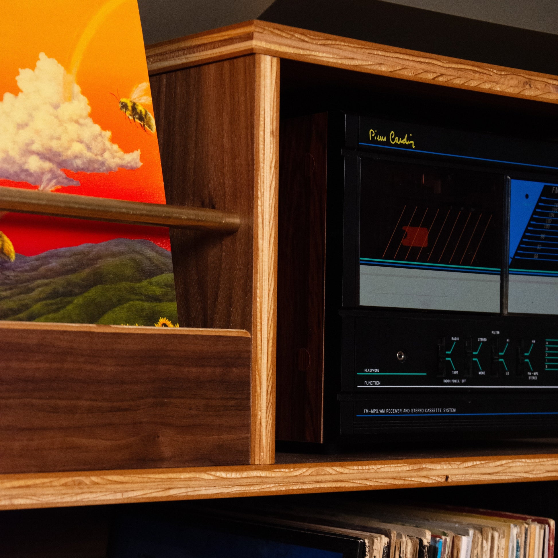 A close-up of a wooden shelf holding a black vintage audio receiver or tape deck with glowing blue and red display elements, alongside vinyl records and a colorful landscape print.