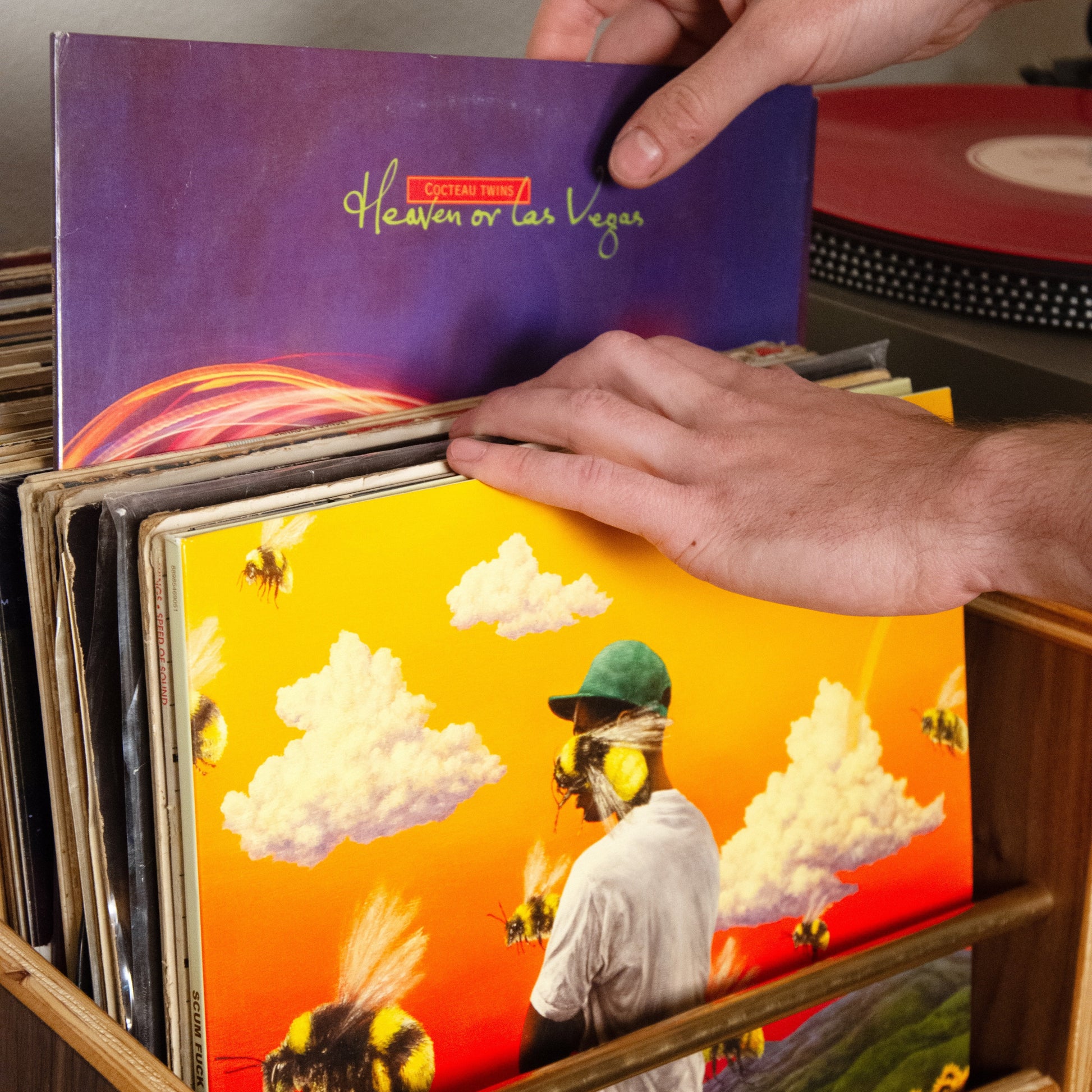A person's hands are shown placing a vinyl record into a wooden record bin. The featured record has a purple cover titled 'Heaven or Hell' with gold lettering, while other records in the bin feature vibrant, colorful album art including a bright yellow and orange scene with a person and a bee.