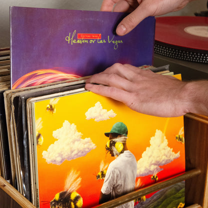 A person's hands are shown placing a vinyl record into a wooden record bin. The featured record has a purple cover titled 'Heaven or Hell' with gold lettering, while other records in the bin feature vibrant, colorful album art including a bright yellow and orange scene with a person and a bee.