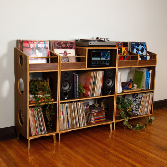 A mid-century modern wooden record shelf unit with open shelves, side cutouts, and small planters, styled with vinyl records, speakers, and equipment against a plain wall on a hardwood floor.
