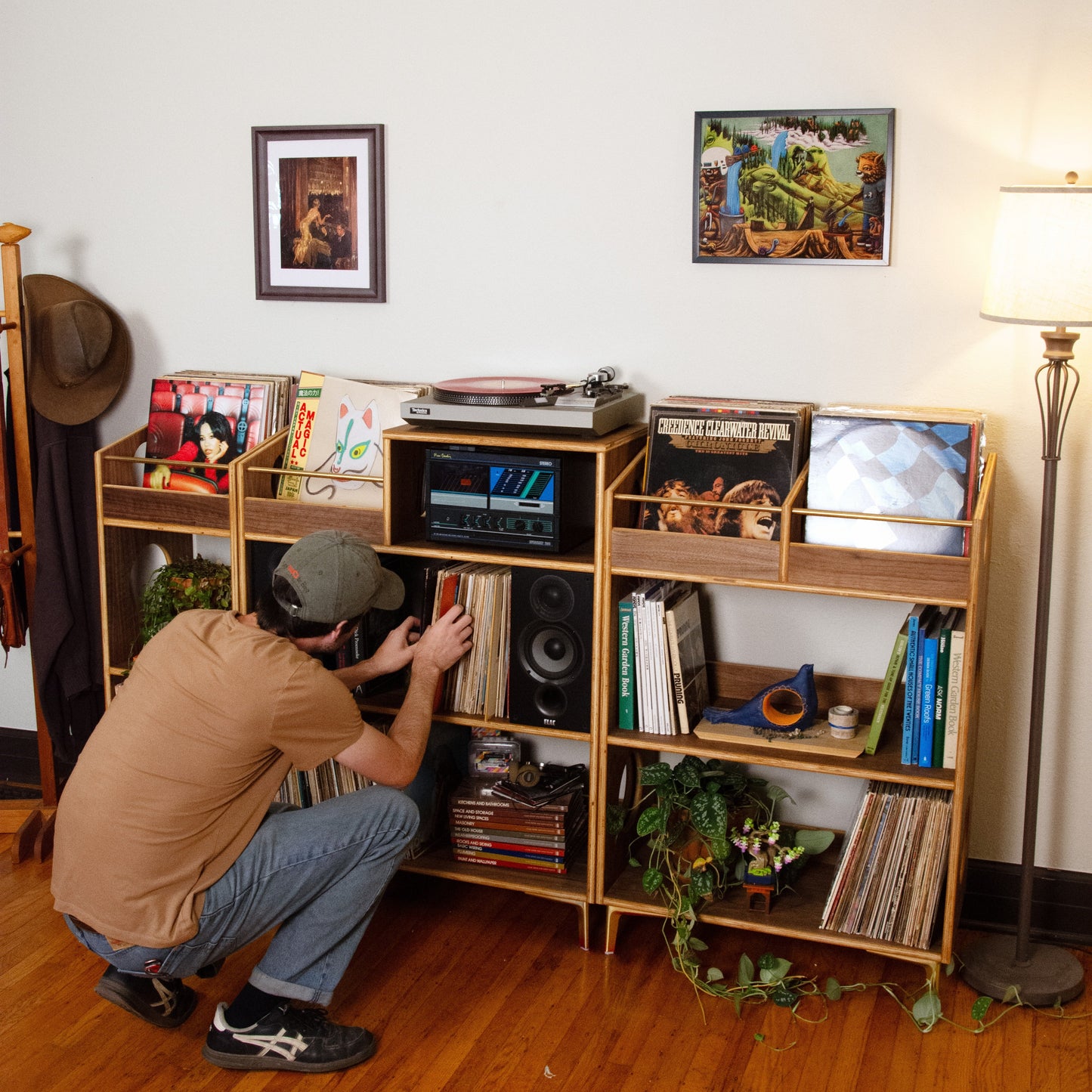 A man in a tan t-shirt and jeans crouches down to arrange records on a wooden multi-shelf bookcase in a room with hardwood floors and a floor lamp.