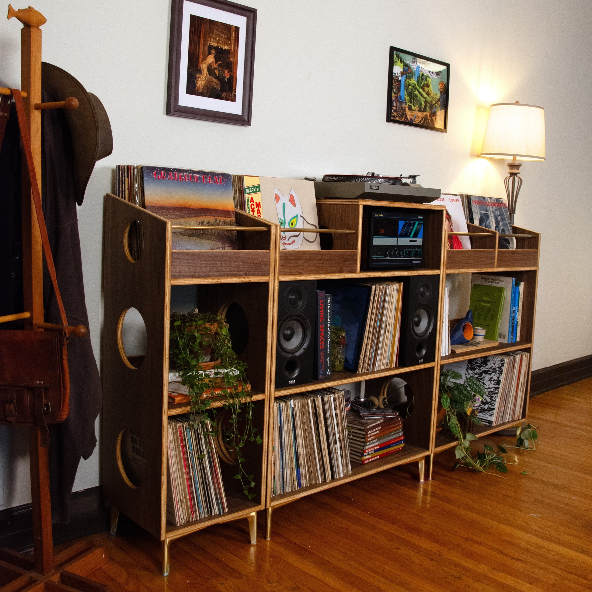 A mid-century modern record rack bookshelf with gold-finished legs and wood-grain fronts, filled with vinyl records, speakers, and plants, set in a room with hardwood floors and a wall lamp.