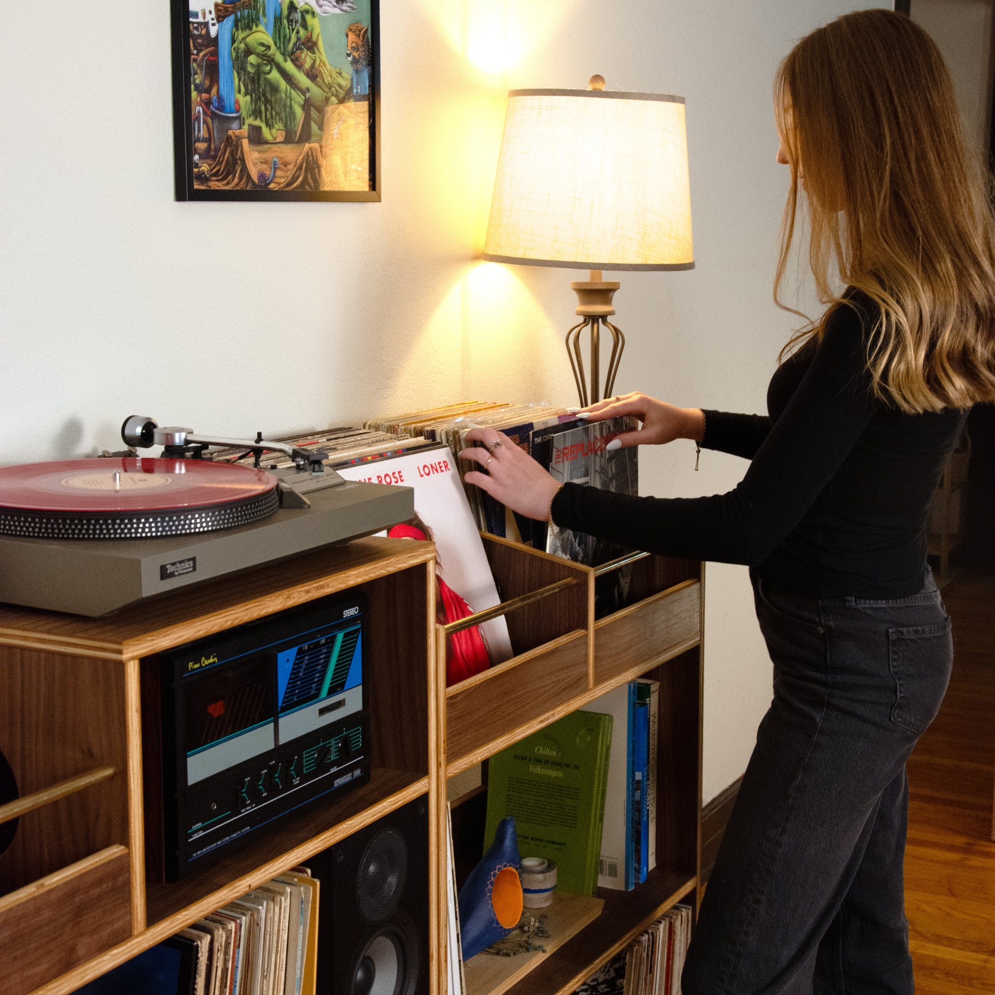 A woman with blonde hair stands at a wooden record shelf, placing a vinyl record next to a turntable and speakers in a warmly lit room.