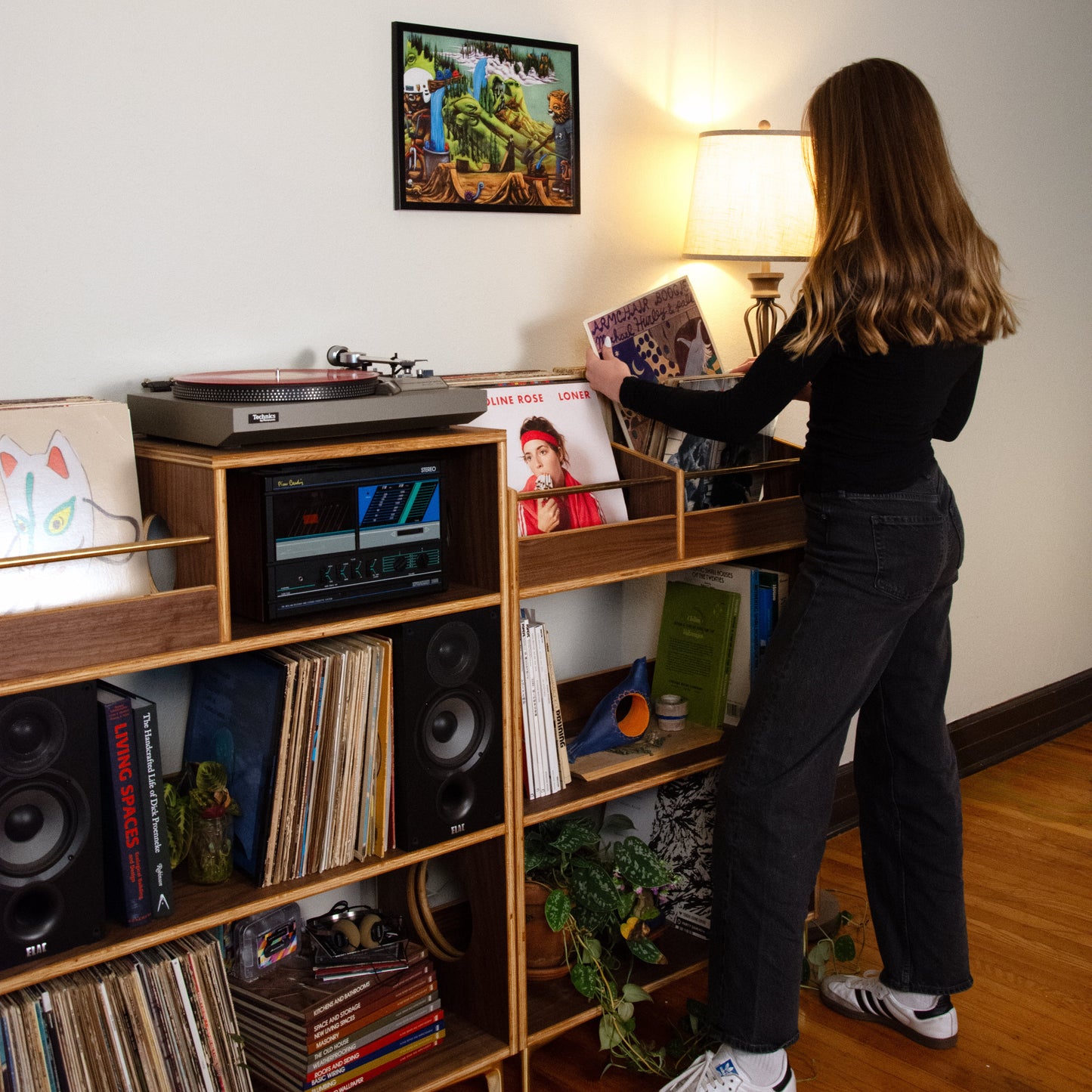 A woman with long brown hair stands in a room, viewed from behind, holding a vinyl record next to a wooden shelf filled with records, speakers, and audio equipment. She is wearing a black long-sleeve top, dark wide-leg jeans, and white sneakers with black stripes.