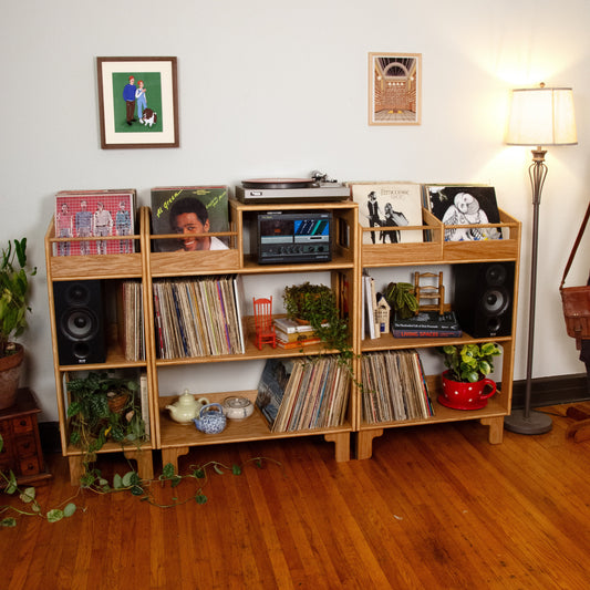 A light natural wood record shelf unit with multiple open cubbies, styled with vinyl records, speakers, framed pictures, plants, and a turntable in a home setting with hardwood floors and a floor lamp.