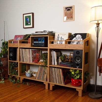 A light natural wood modular record shelf unit styled with vinyl records, speakers, a turntable, and small decor items in a room with hardwood floors and a floor lamp.