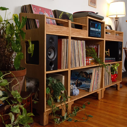 A tall, light wood record shelf unit styled in a cozy room corner with plants, speakers, and vinyl records, next to a lamp and coat rack.