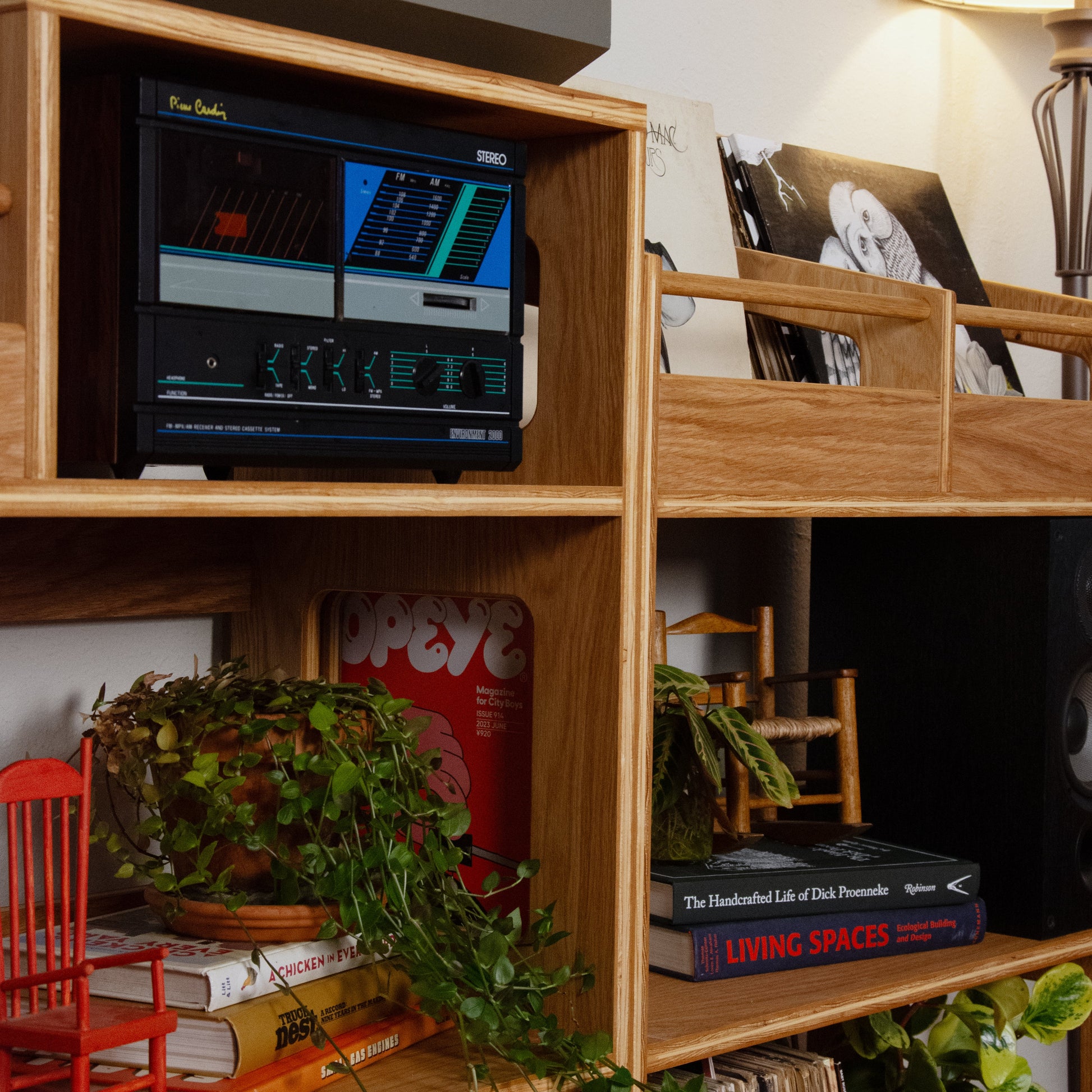 A wooden shelving unit in a cozy room setting, styled with a turntable, books, plants, a red chair figurine, and vinyl records. A small black electronic device with blue and green display screens is mounted on one of the shelves.