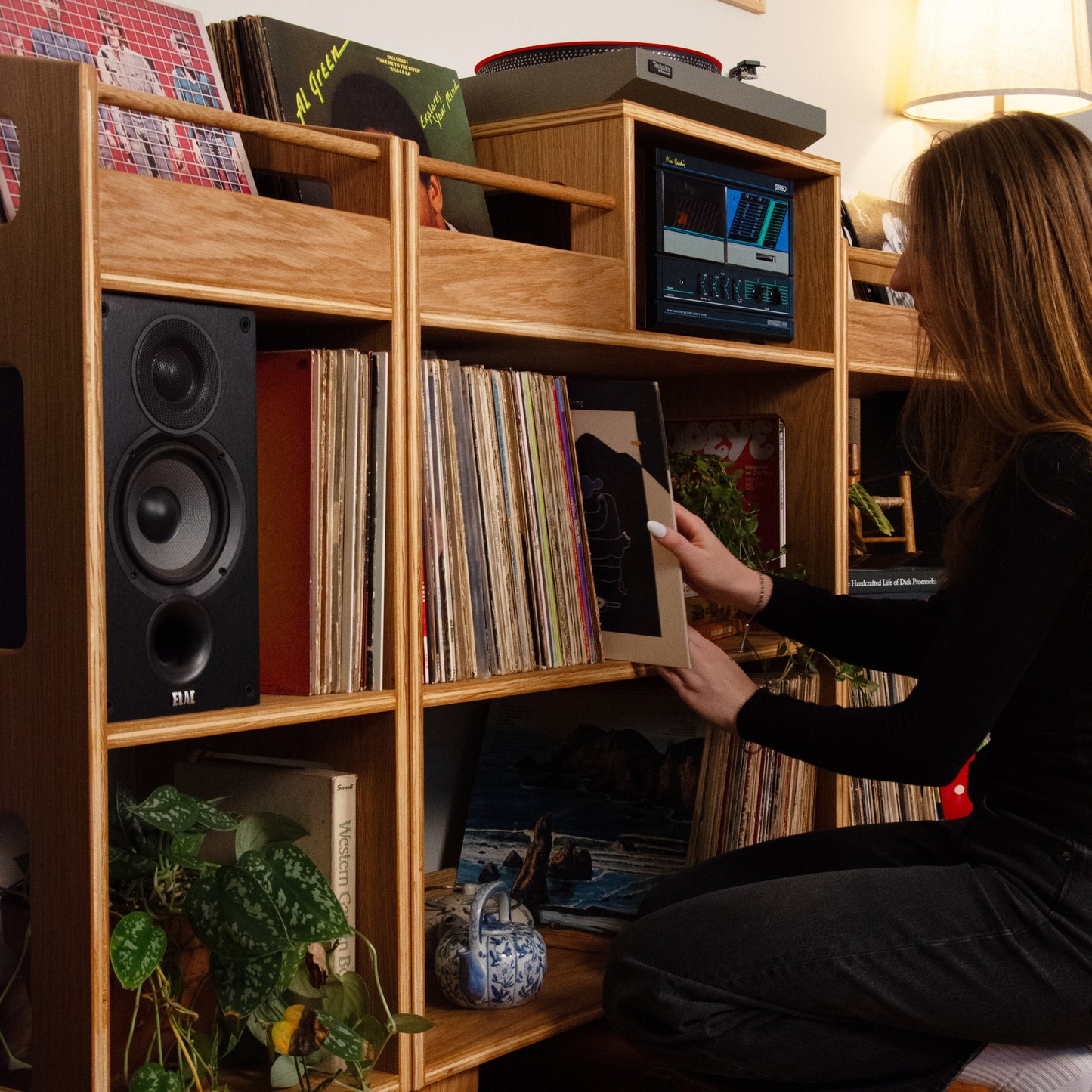 A woman kneels beside a large wooden record shelf in a cozy room, holding a vinyl record. The shelf is filled with vinyl records, speakers, a turntable, and plants, with a lamp glowing in the background.