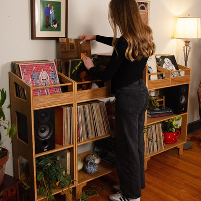 A woman with long light brown hair stands in a room, looking into a wooden record shelf filled with vinyl records, speakers, framed pictures, and plants. The room has a warm, cozy atmosphere with a lit table lamp on the right and hardwood floors.