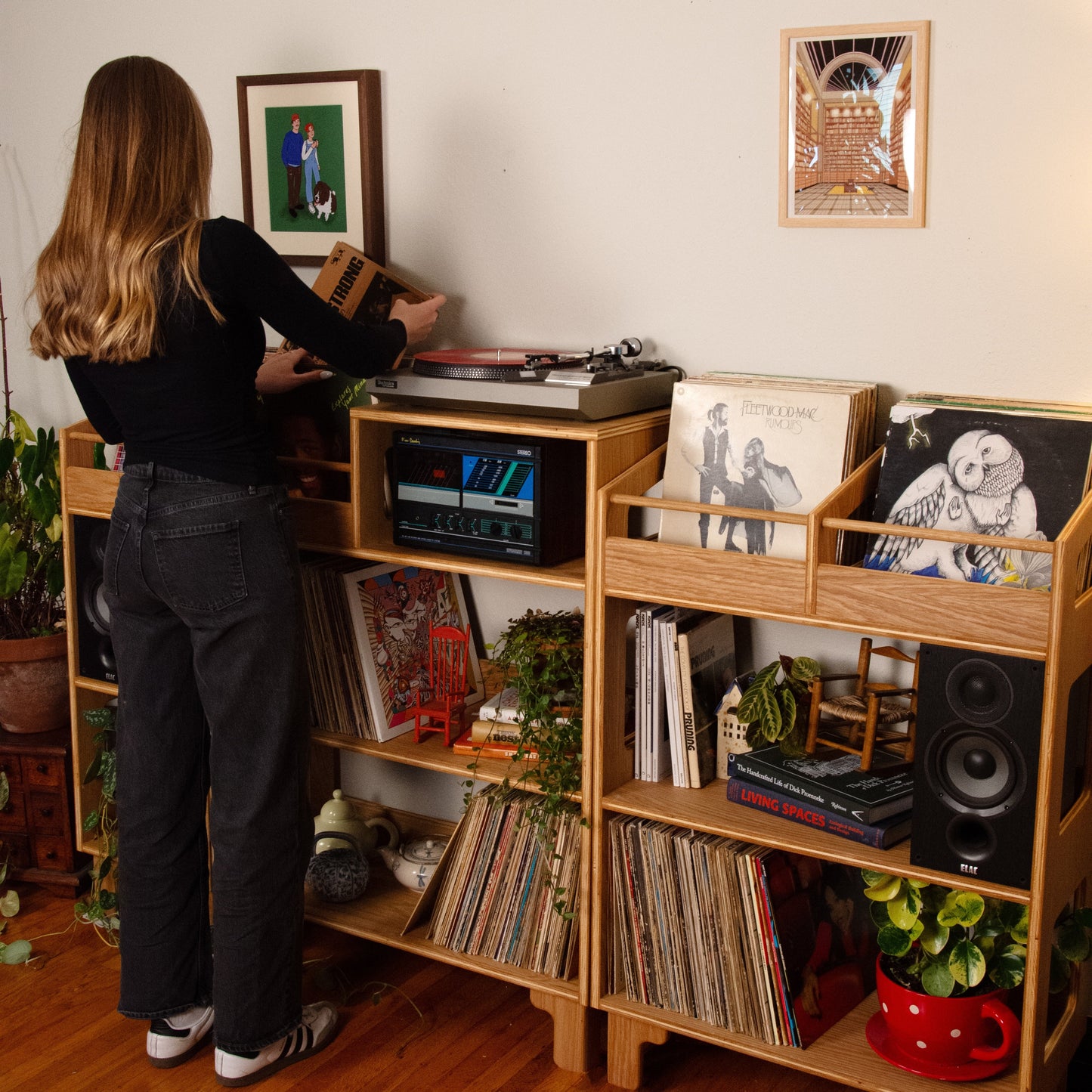 A woman stands at a wooden record shelf in a room with hardwood floors, adjusting a turntable. The shelf is filled with vinyl records, books, plants, and framed wall art.