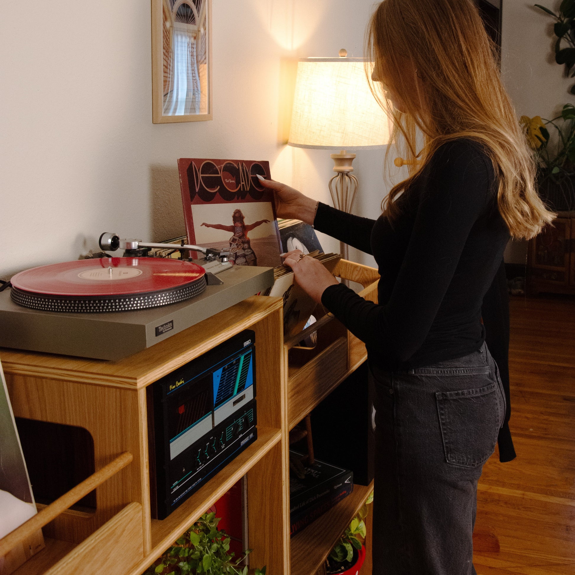 A woman with long blonde hair stands in a room next to a wooden record stand, holding a vinyl record in front of a glowing lamp. A turntable with a pink vinyl record sits on the stand, which also holds a cassette player.
