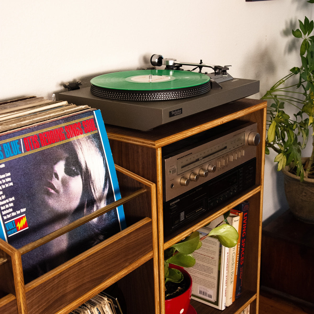 A wooden record shelf in a home setting, holding a turntable with a green vinyl record, a stereo receiver, and stacks of vinyl records.
