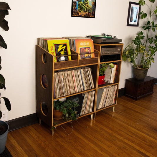 A mid-century modern wooden record shelf with circular cutouts, holding vinyl records, books, a turntable, and small plants, set in a room with hardwood floors and white walls.