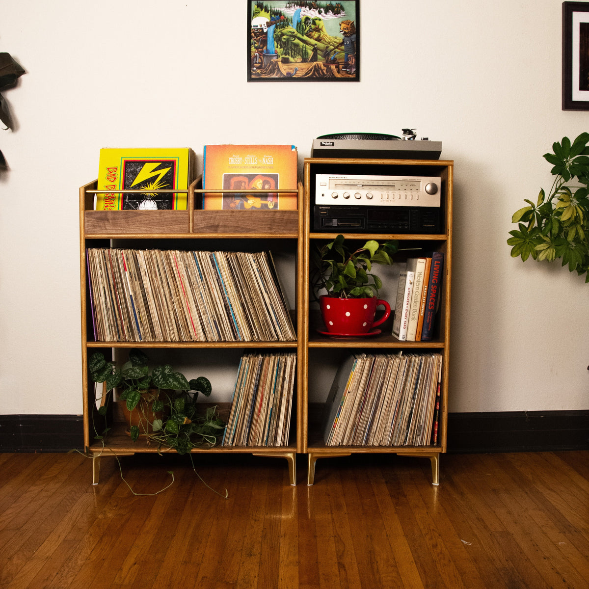 A two-tier wooden vinyl record shelf with gold-toned metal legs, filled with records, books, a turntable, and small potted plants, set against a white wall on a hardwood floor.
