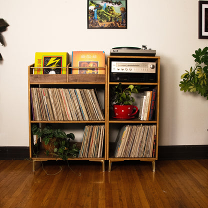 A two-tier wooden vinyl record shelf with gold-toned metal legs, filled with records, books, a turntable, and small potted plants, set against a white wall on a hardwood floor.