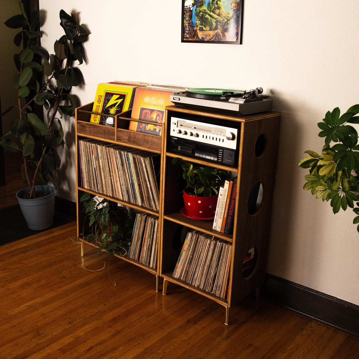 A mid-century modern wooden record shelf filled with vinyl records, holding a turntable and audio equipment, set in a room with hardwood floors, houseplants, and a framed picture on the wall.