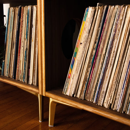 A close-up of a mid-century modern wooden record shelf filled with vinyl records, featuring a red ceramic mug on the top shelf and a polished wood floor below.