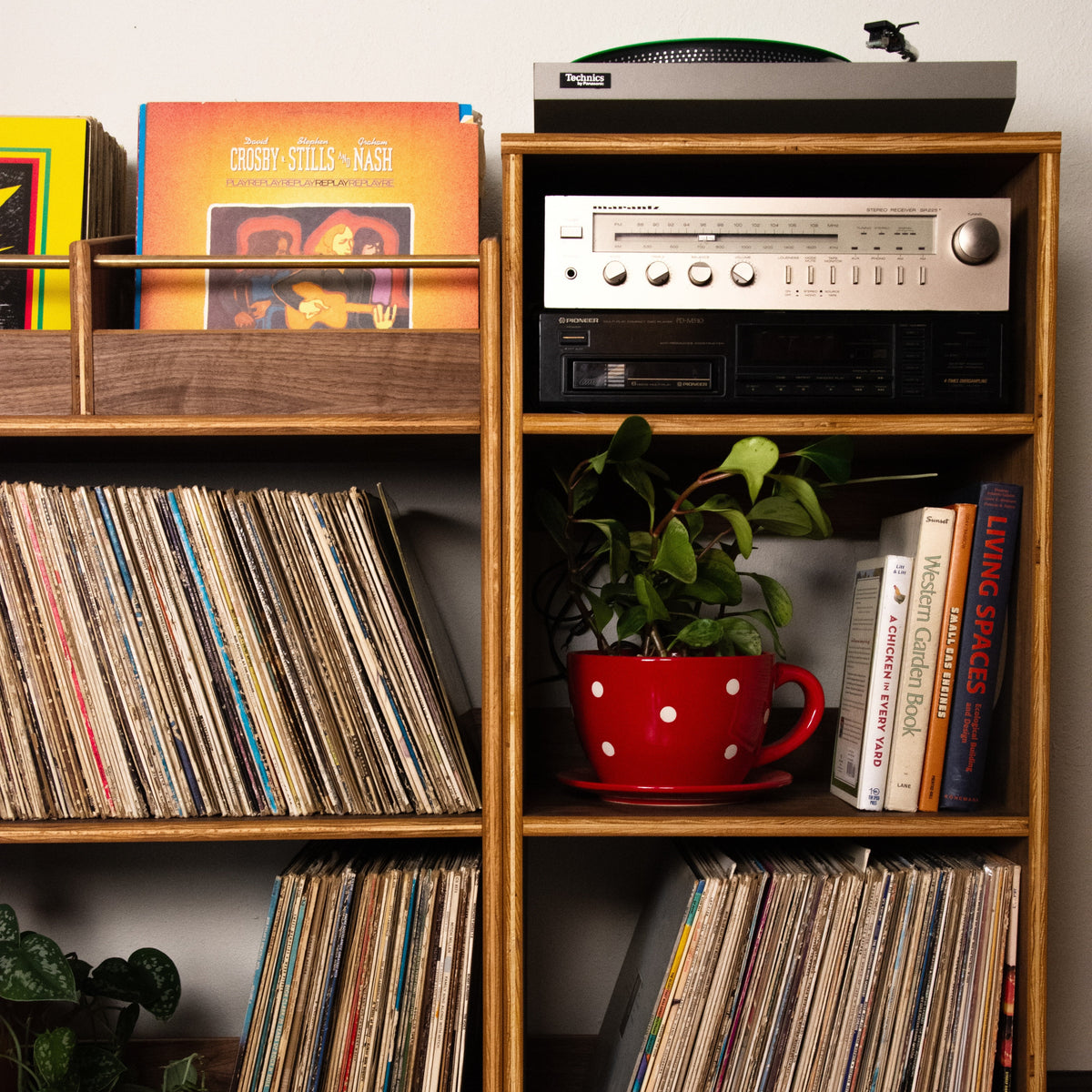 A mid-century modern wooden record shelf with gold-toned legs, filled with vinyl records, books, a potted plant in a red polka-dot mug, and a turntable on top, set against a plain wall and hardwood floor.