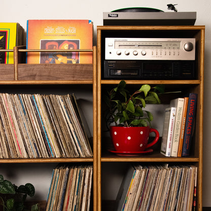 A mid-century modern wooden record shelf with gold-toned legs, filled with vinyl records, books, a potted plant in a red polka-dot mug, and a turntable on top, set against a plain wall and hardwood floor.