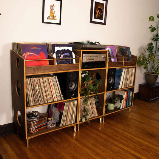 A mid-century modern wooden record shelf with gold legs and circular cutout details, filled with vinyl records, speakers, and audio equipment, set in a room with hardwood floors and framed wall art.