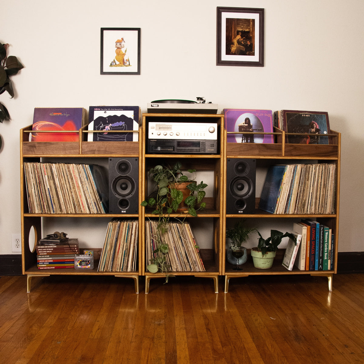 A mid-century modern wooden record shelf with gold-tone legs and brackets, filled with vinyl records, speakers, a turntable, and small potted plants, set against a plain beige wall with framed art on a hardwood floor.