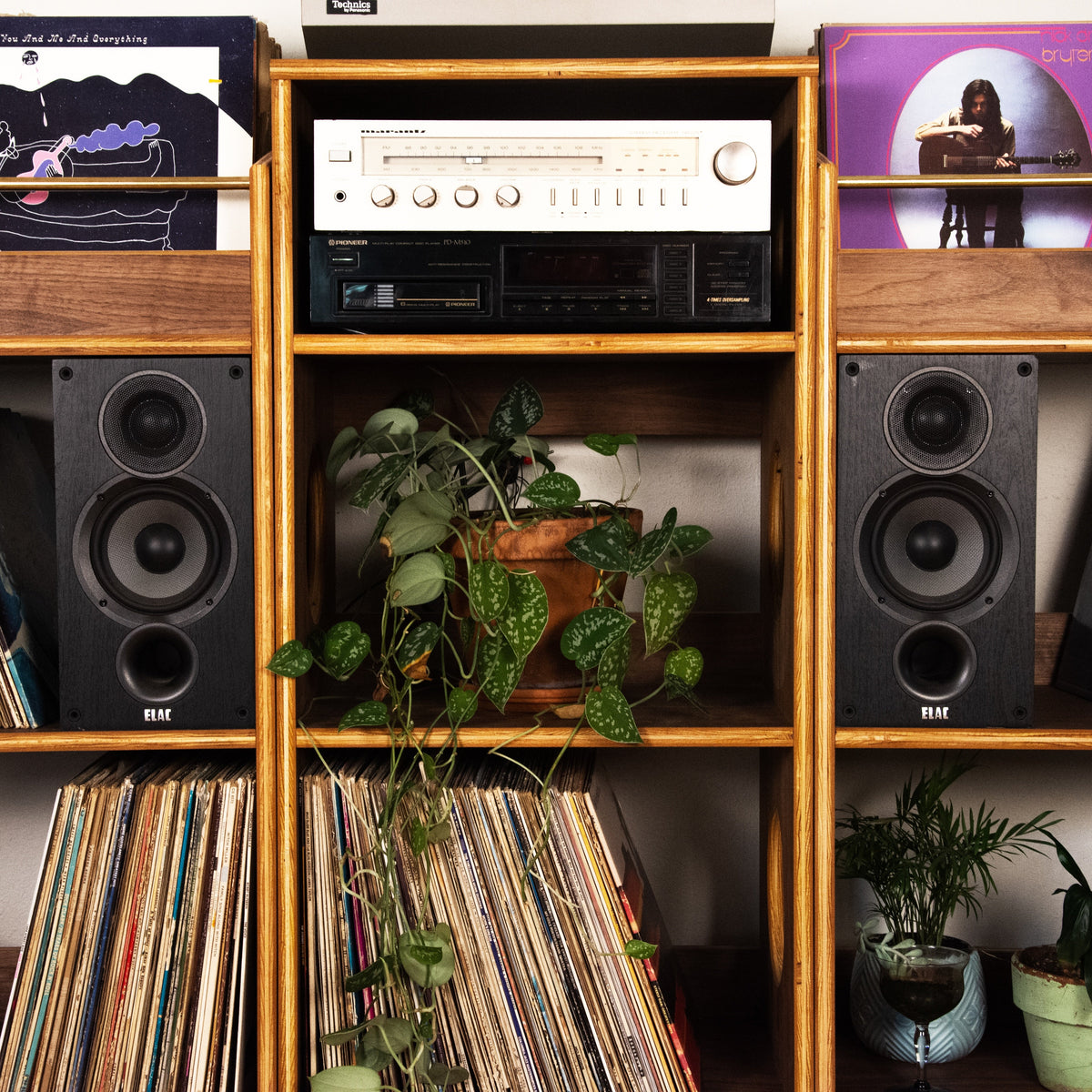 A wooden record shelf filled with vinyl records, two black speakers, a vintage stereo receiver, and green potted plants.