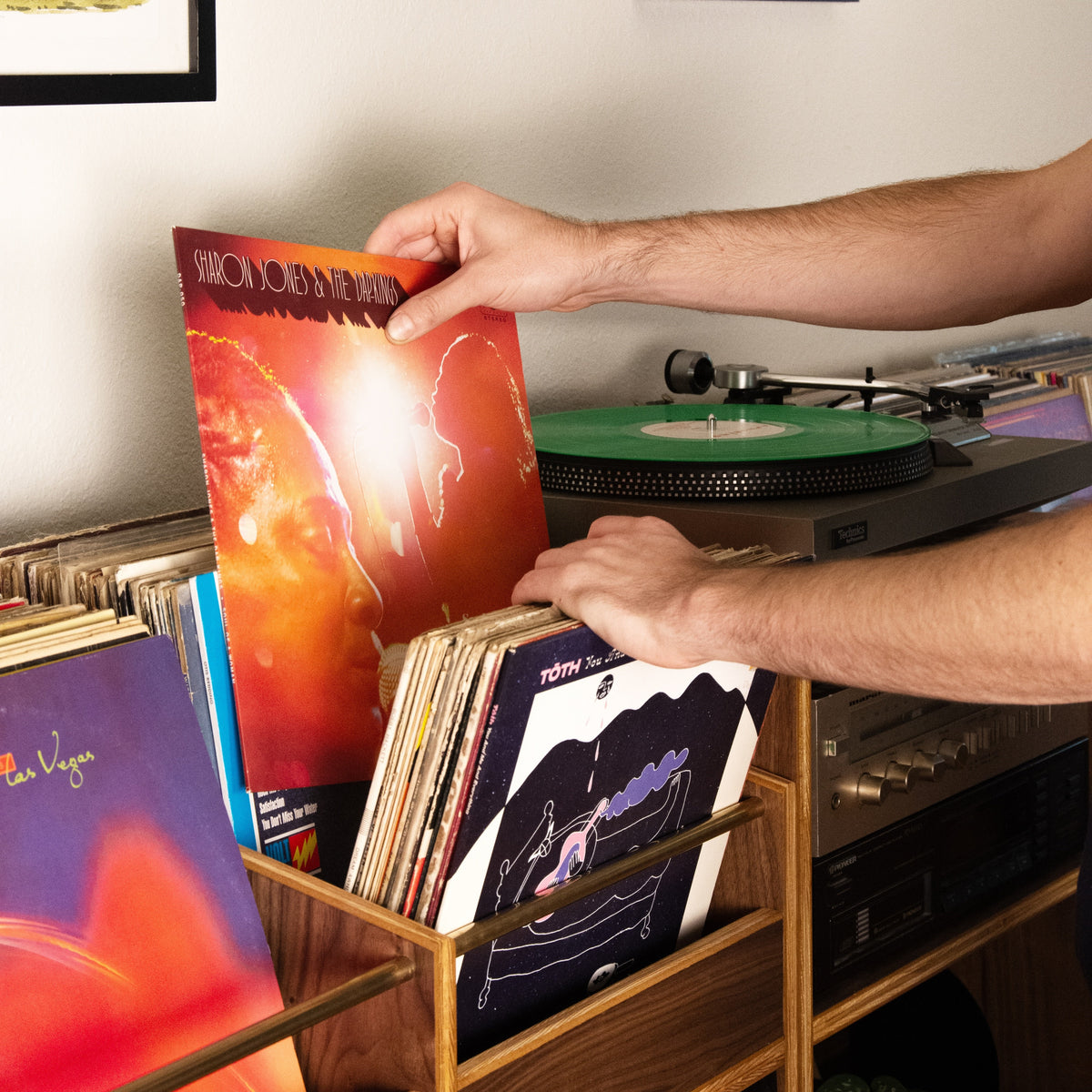 A person's hands holding a vinyl record sleeve with a fiery red and orange cover, placing it into a wooden record rack next to a turntable in a room setting.