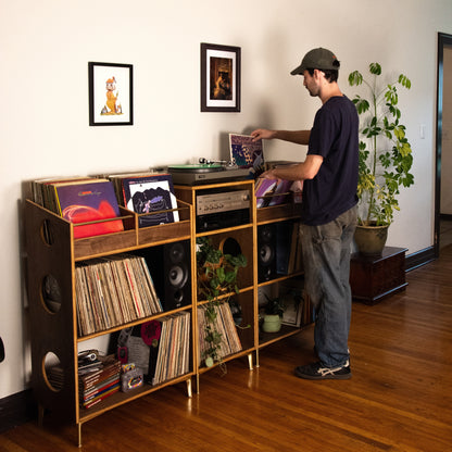 A man in a dark cap and navy t-shirt stands next to a large wooden record rack filled with vinyl records and speakers in a home interior with hardwood floors and a potted plant.