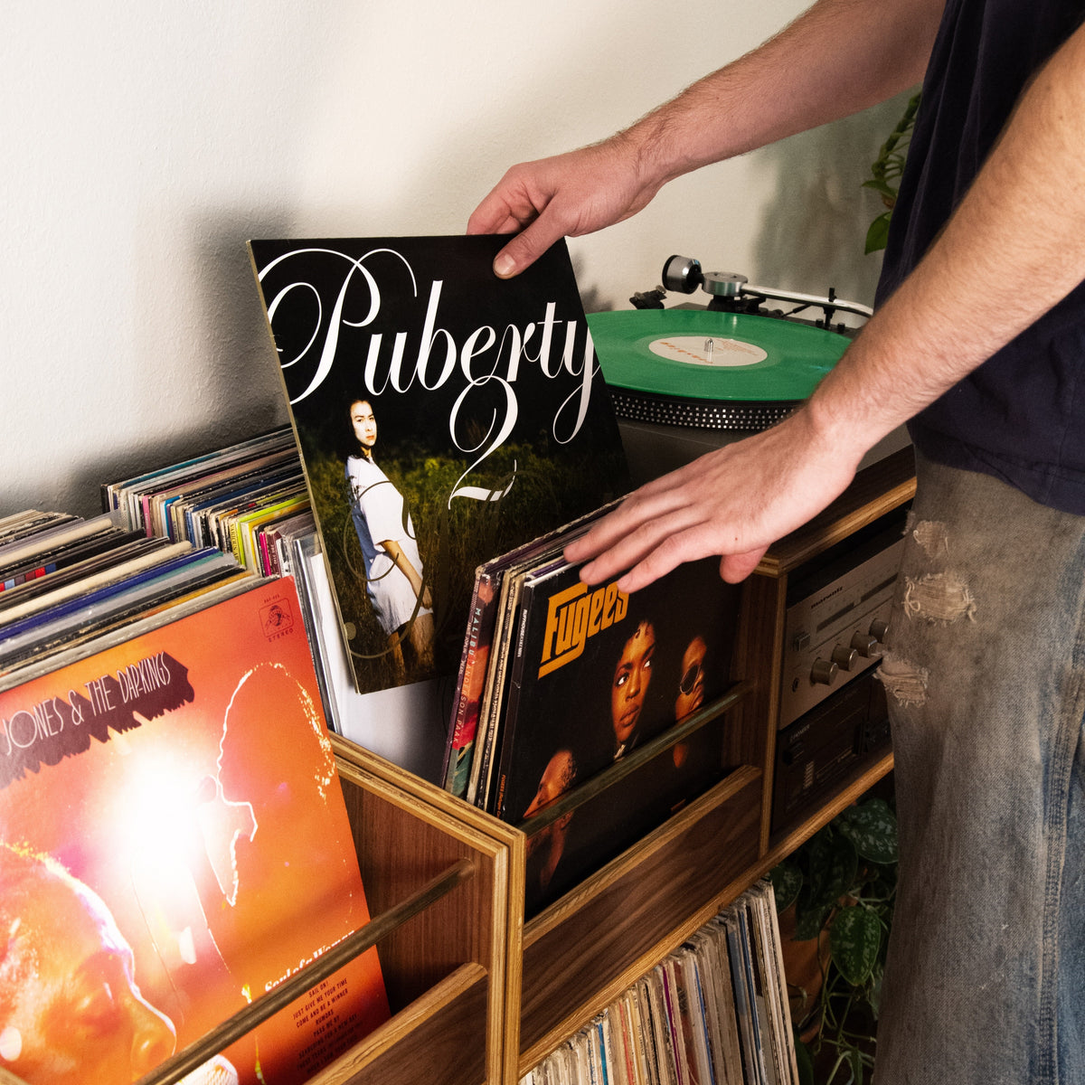 A person's hands and torso are shown selecting a vinyl record from a wooden record bin filled with records. The person is holding a black vinyl record with 'Puberty' written on the cover.