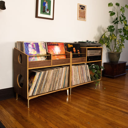 A mid-century modern wooden record shelf with gold-toned metal legs, filled with vinyl records, in a room with hardwood floors, a white wall, and a potted plant.