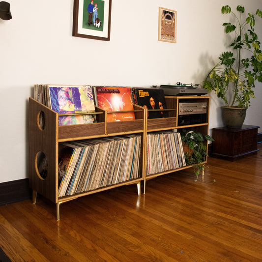 A mid-century modern wooden record shelf with gold-toned metal legs, filled with vinyl records, in a room with hardwood floors, a white wall, and a potted plant.