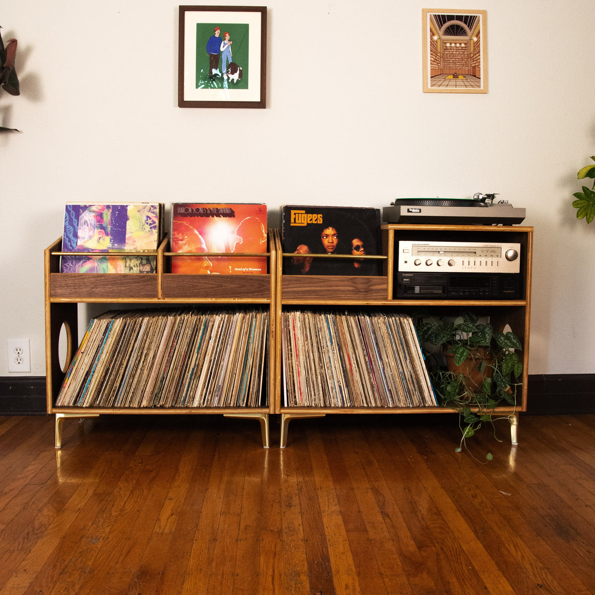 A mid-century modern wooden record rack with gold-toned legs, filled with vinyl records and topped with a turntable and receiver, set against a white wall with framed art on a hardwood floor.