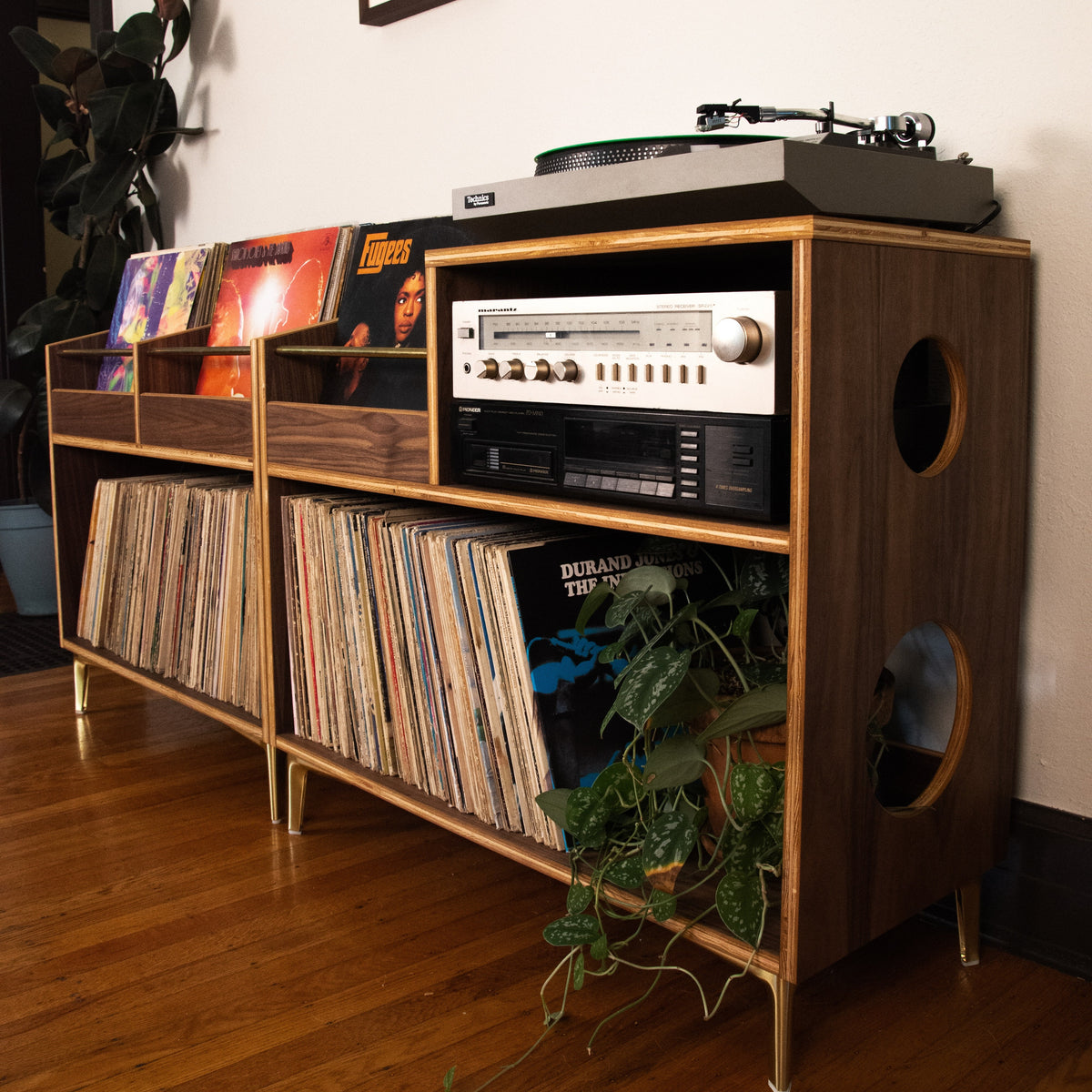 A mid-century modern wooden record rack with gold-toned legs and circular cutouts, styled in a living room with vinyl records, a turntable, and a plant on a hardwood floor.