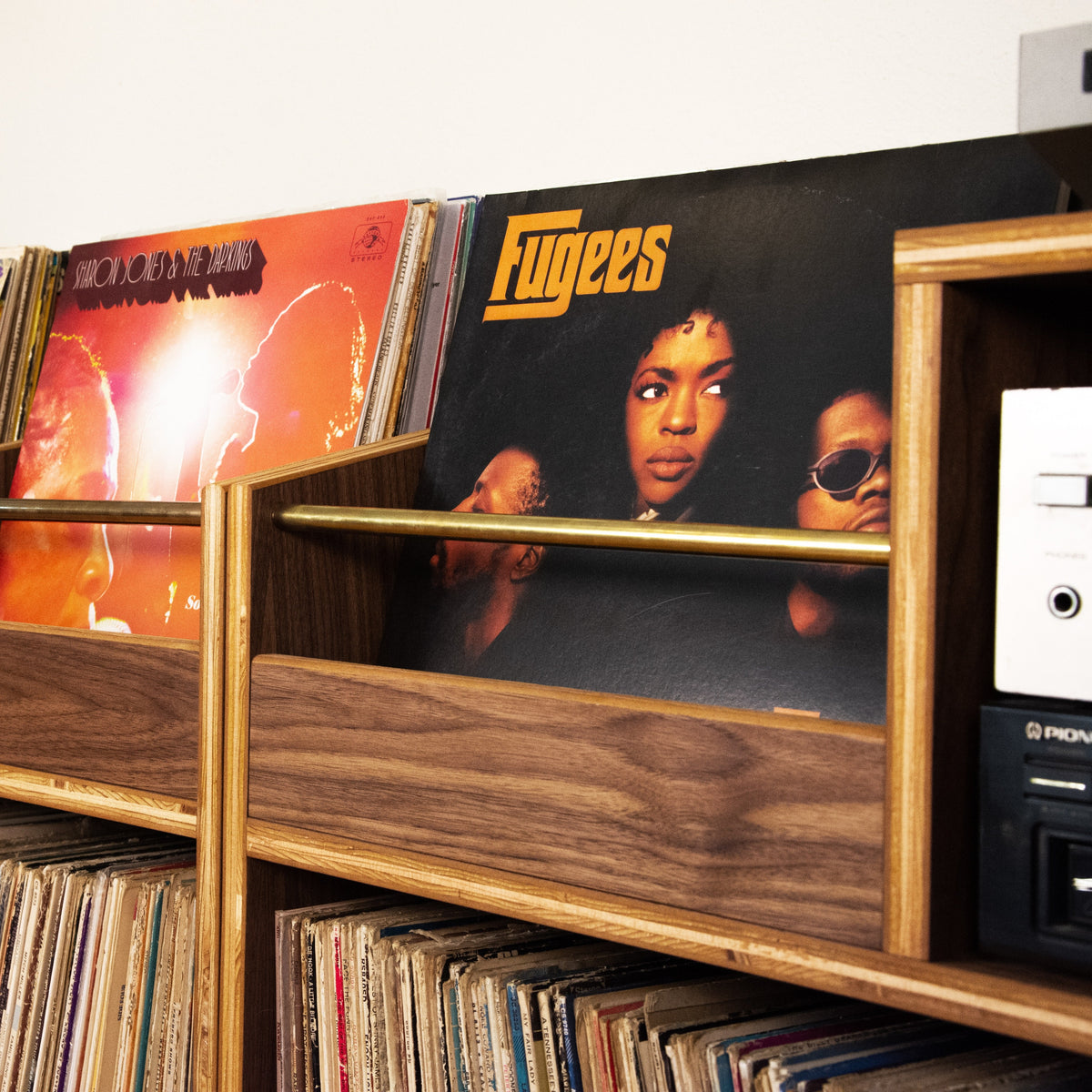 A wooden record shelf holding Fugees vinyl records, with other records and a Pioneer cassette player visible in a home or studio setting.