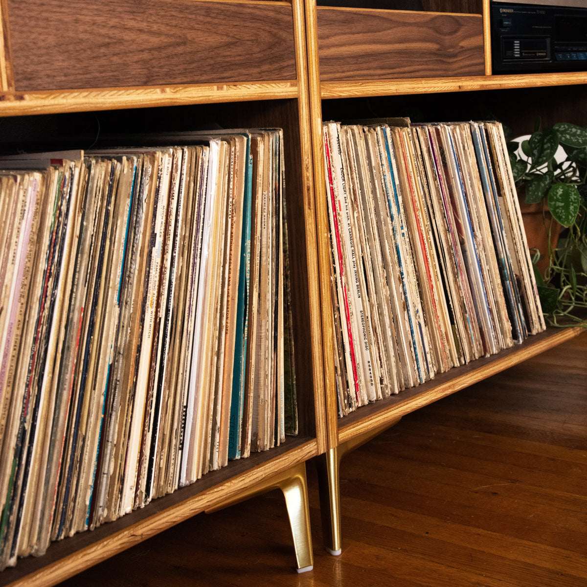 A mid-century modern wooden record shelf filled with vinyl records, with record sleeves visible on the upper shelves and a stereo receiver on the side.