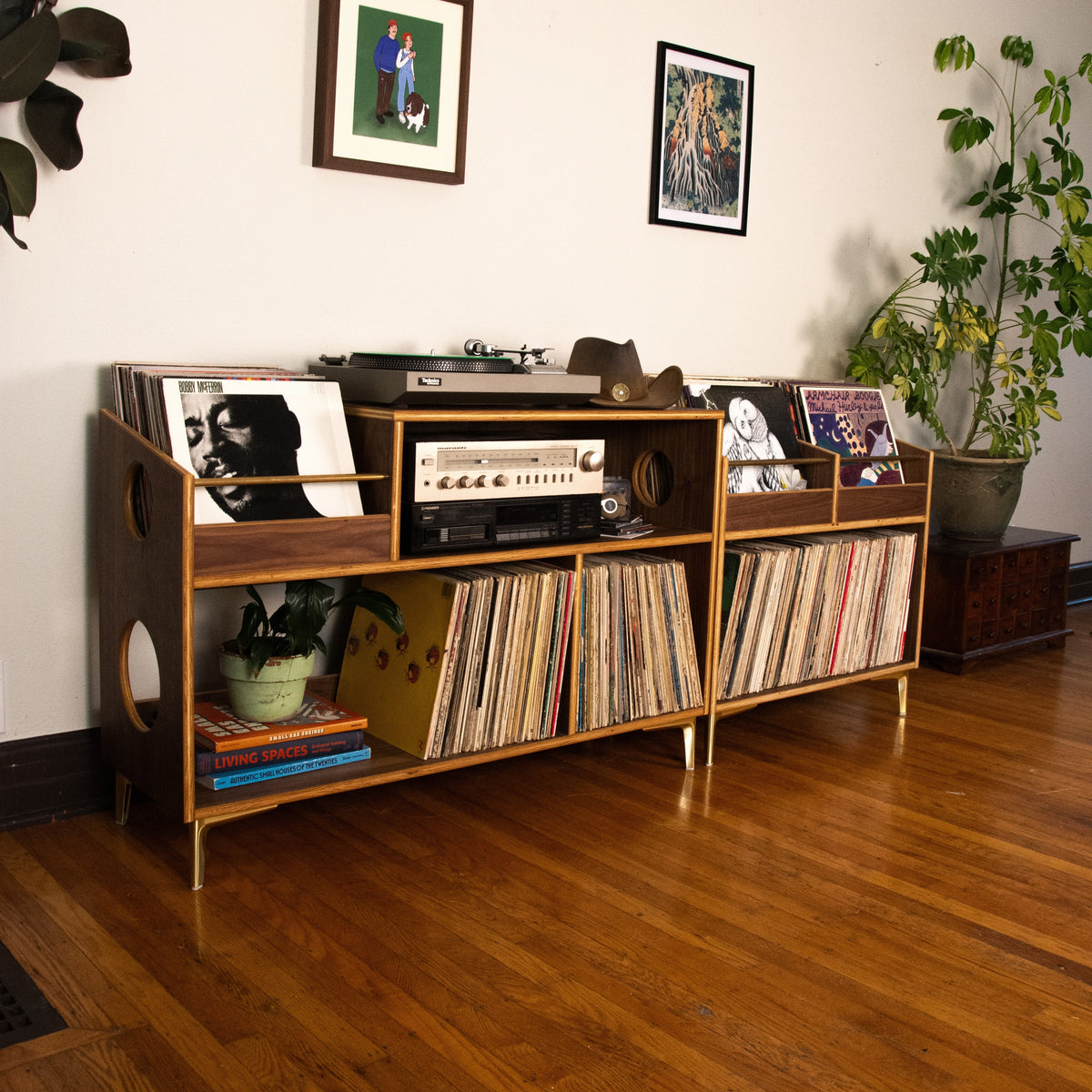 A mid-century modern wooden record shelf with gold-tone legs, styled in a living room with vinyl records, a turntable, framed wall art, and potted plants.