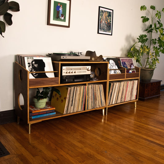 A mid-century modern wooden record shelf with gold-tone legs, styled in a living room with vinyl records, a turntable, framed wall art, and potted plants.