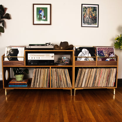 A mid-century modern wooden record shelf with gold-toned legs and trim, styled with vinyl records, a turntable, and framed art on a white wall above a hardwood floor.