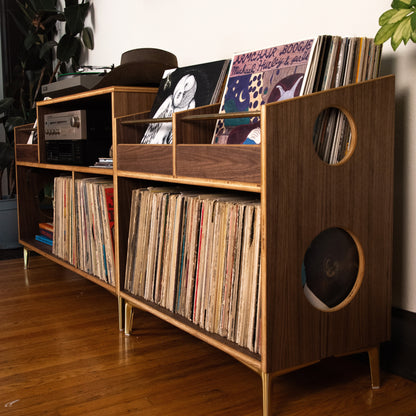 A mid-century modern wooden record shelf with gold-toned legs and circular cutouts, filled with vinyl records in a home interior setting.