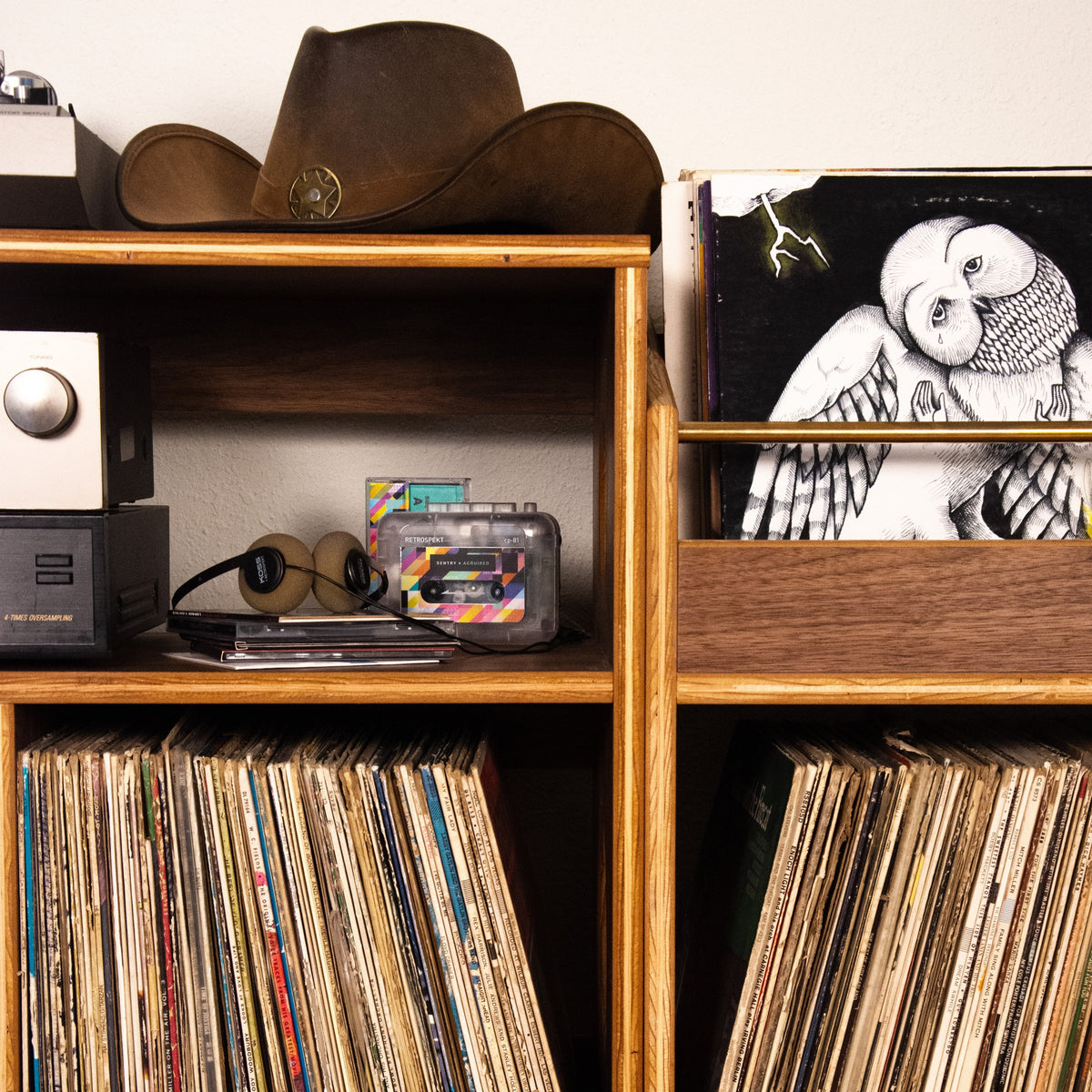 A pair of mid-century modern wooden record shelves filled with vinyl records, featuring a brown cowboy hat and a record sleeve with an owl illustration on top.
