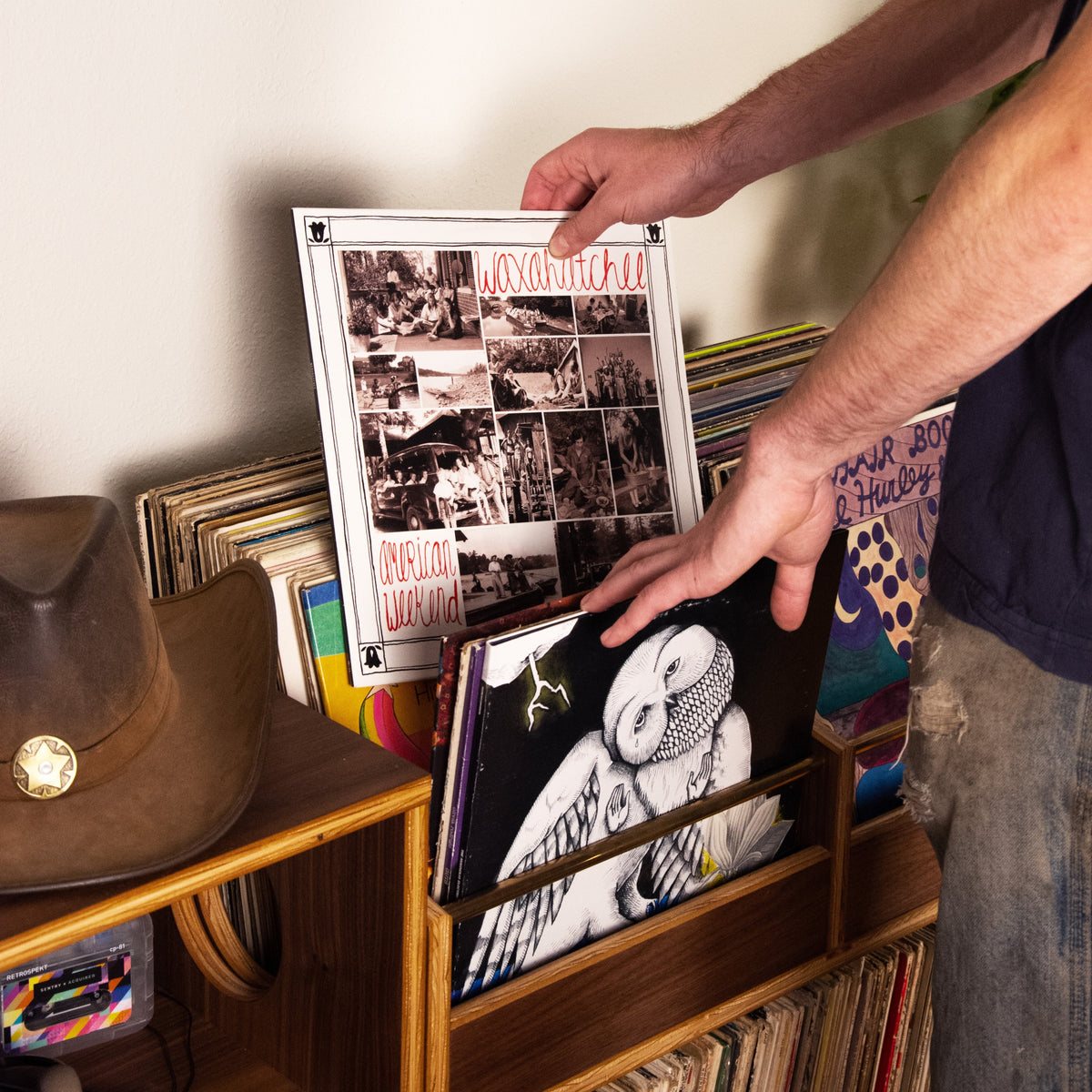 A person's hands holding a framed black-and-white album cover in a wooden record rack filled with vinyl records, with a framed art print on the wall above.