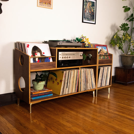 A mid-century modern wooden record player console cabinet with gold legs, styled in a living room with vinyl records, a turntable, and plants on a hardwood floor.