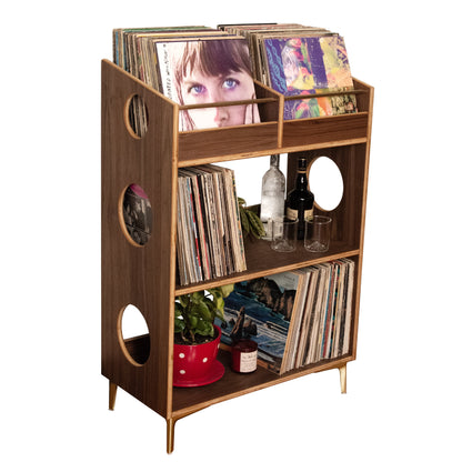 A mid-century modern wooden record shelf with open cubbies, circular cutouts, and tapered legs, styled with vinyl records, bottles, and small plants against a solid white background.