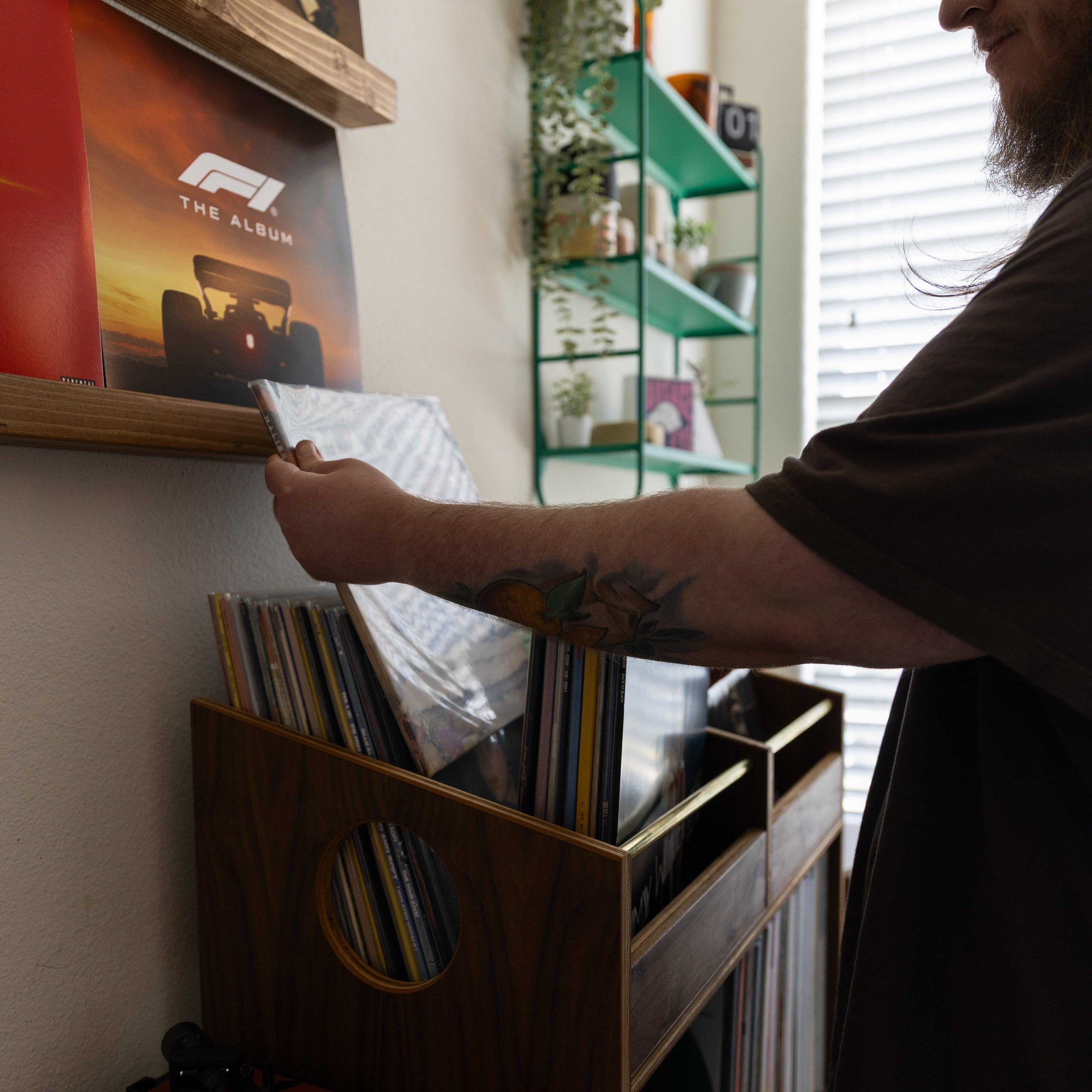 A man with glasses and a beard is seen from the side, reaching for a vinyl record inside a wooden record organizer on a shelf in a home interior.